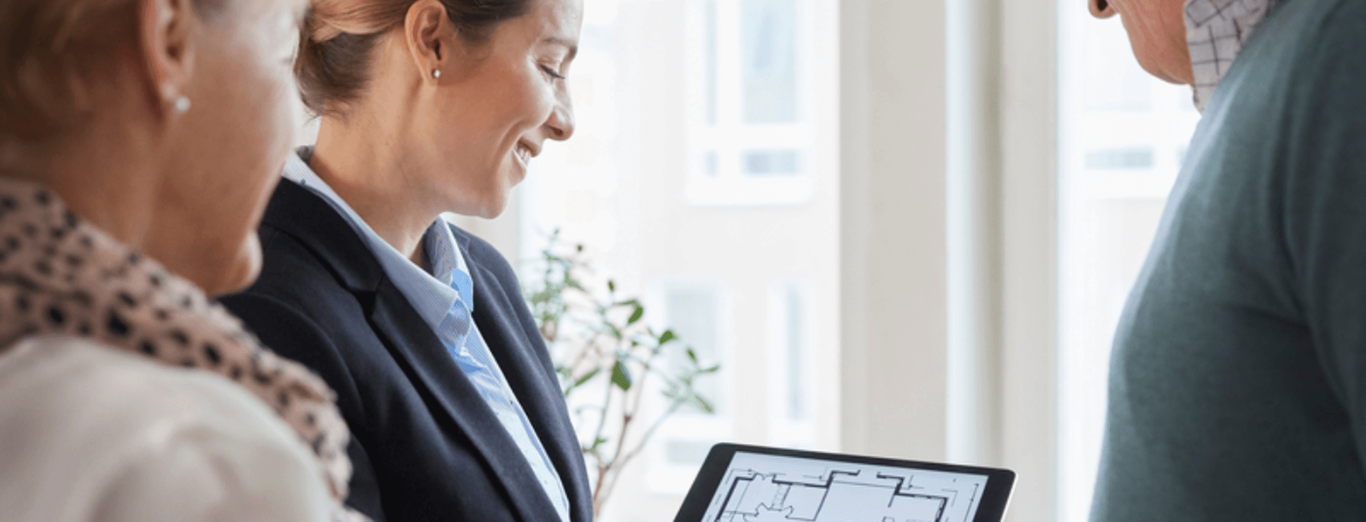 Un agent immobiliari mostra un plànol en una tauleta a una parella de gent gran. L'agent està somrient.