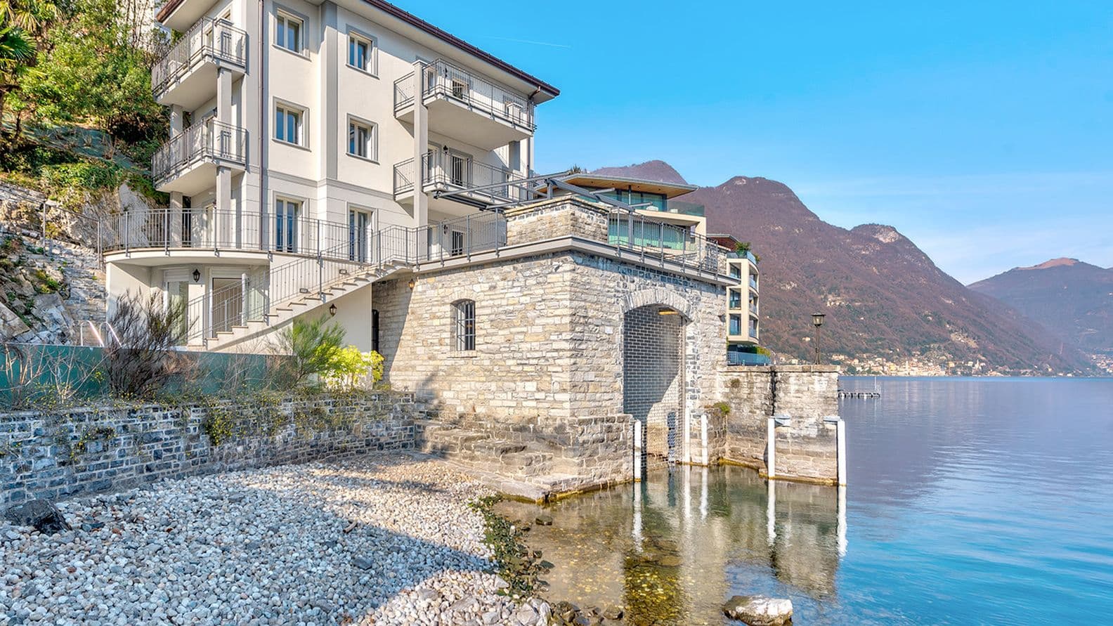 Villa am Seeufer mit Steinfassade, mehreren Balkonen und einem kleinen Bootssteg vor der Kulisse der Berge unter einem klaren blauen Himmel. Im Vordergrund die felsige Küste.