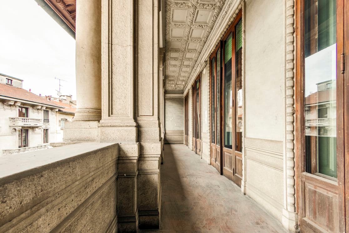 Ein langer, schmaler Balkon mit Steinsäulen und Holztüren mit Blick auf eine Stadtlandschaft mit roten Ziegeldächern unter einem bewölkten Himmel.