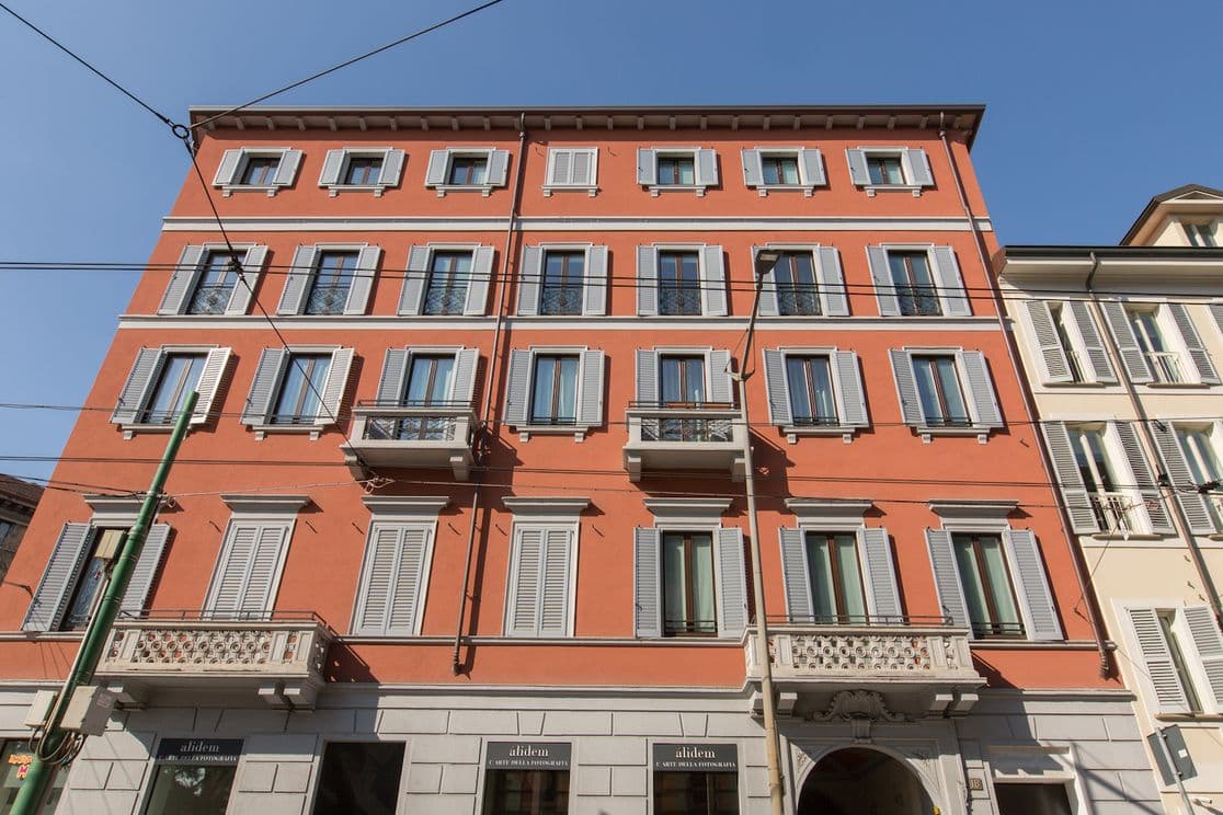 Ein vierstöckiges rotes Gebäude mit grauen Fensterläden und Balkonen vor einem klaren blauen Himmel. Im Vordergrund sind Straßenbahnkabel zu sehen.