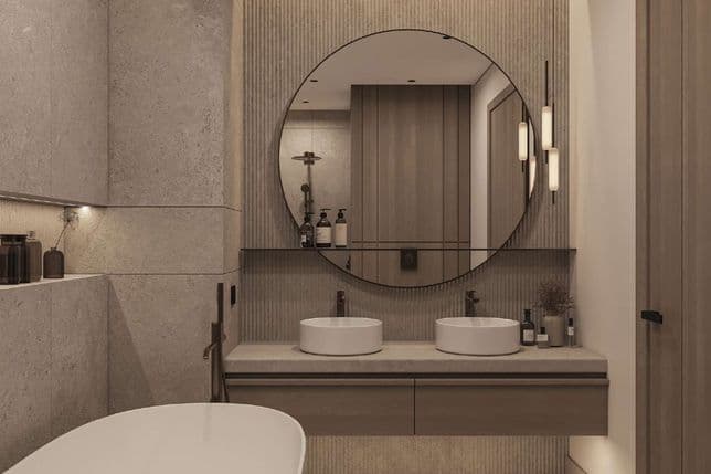 Modern bathroom with a freestanding tub, double sinks, round mirror, and grey stone tiles, featuring soft ambient lighting.