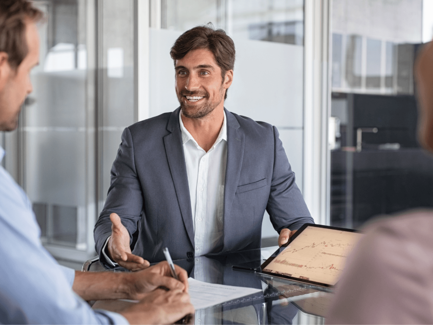 Un home somrient amb un vestit gris presenta una tauleta amb un gràfic a uns clients en una taula de vidre. Un client signa un document.