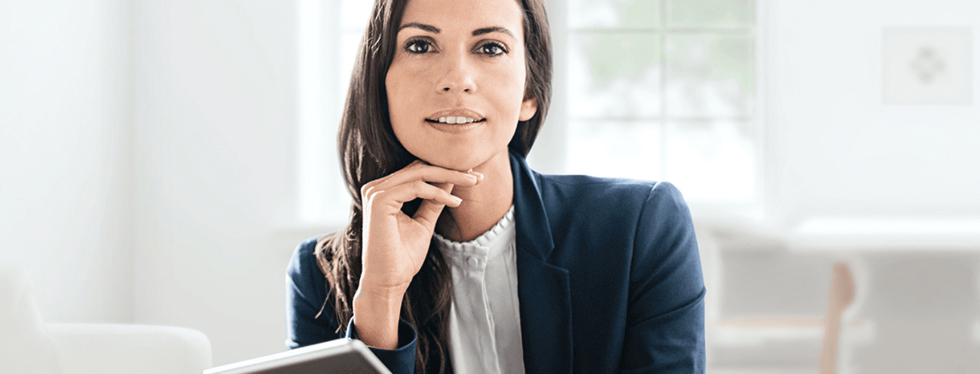 A woman in a blue blazer holds a tablet, resting her chin on her hand in a bright, modern room.