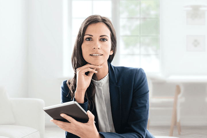 Una dona amb una americana blava sosté una tauleta, recolzant la barbeta a la mà en una habitació moderna i lluminosa.