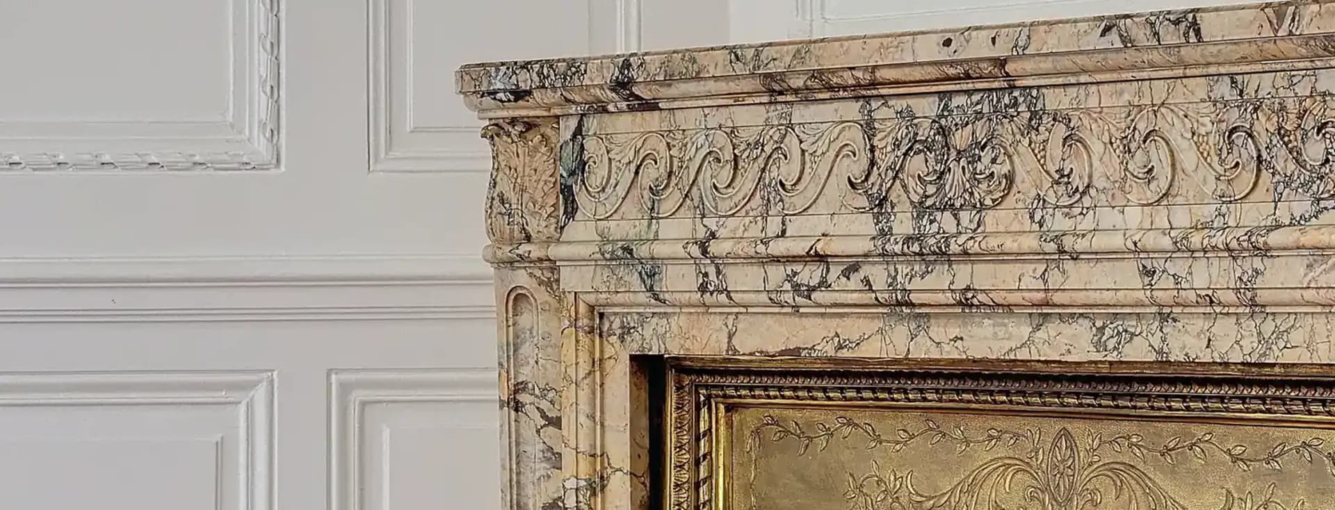Close-up van een marmeren open haard met gouden en zwarte accenten tegen een wit lambrisering en een glimp van een spiegel erboven.