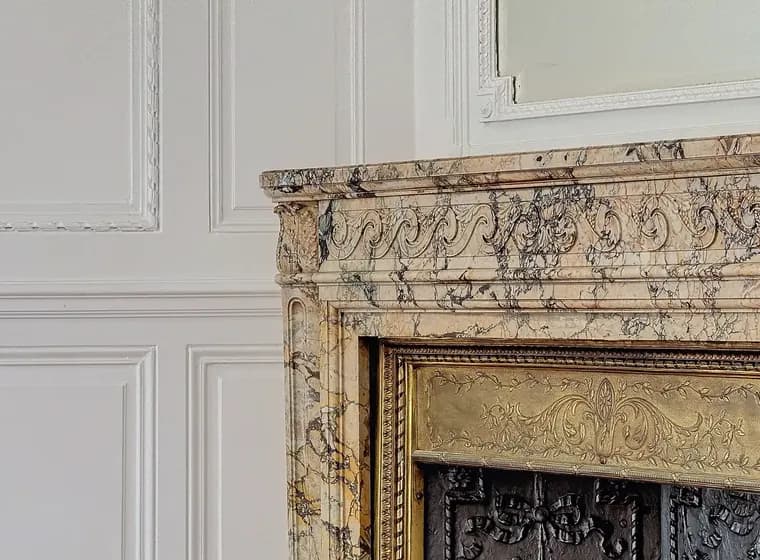 Close-up van een marmeren open haard met gouden en zwarte accenten tegen een wit lambrisering en een glimp van een spiegel erboven.