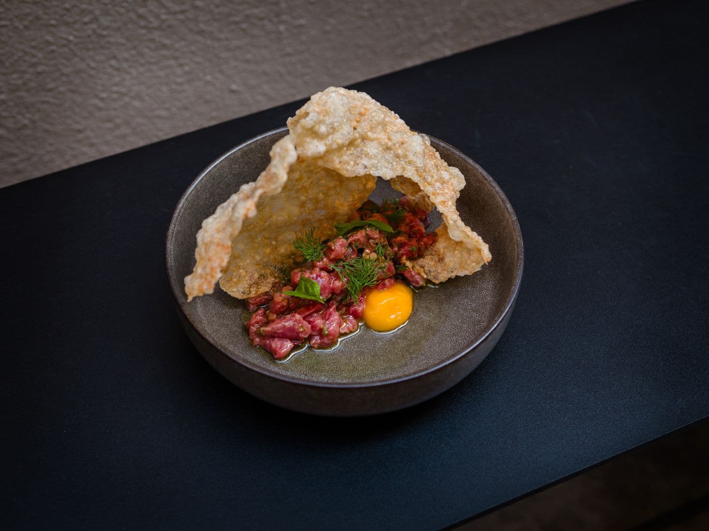 Ένα γκρι μπολ περιέχει ταρτάρ βοδινού με έναν έντονα κίτρινο κρόκο αυγού, γαρνιρισμένο με άνηθο και ένα μεγάλο, τραγανό κράκερ.