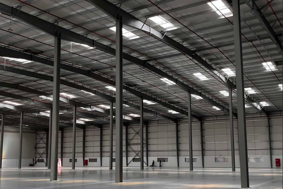 Spacious, empty warehouse interior with high ceilings, metal beams, and concrete floor, illuminated by skylights and ceiling lights.
