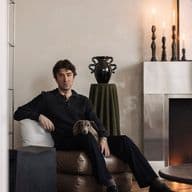 Man seated on a brown cushion, with a small dog in his lap, in a modern living room with a decorated mantle and candlesticks.