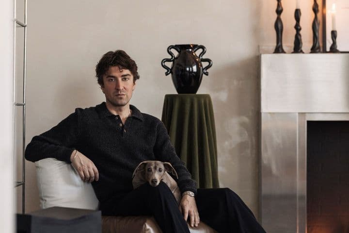 Man seated on a brown cushion, with a small dog in his lap, in a modern living room with a decorated mantle and candlesticks.