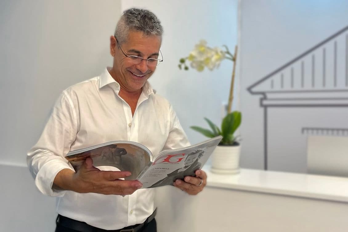 A man in a white shirt reads a magazine, standing next to a reception desk with "ENGEL & VÖLKERS" logo and a potted orchid.