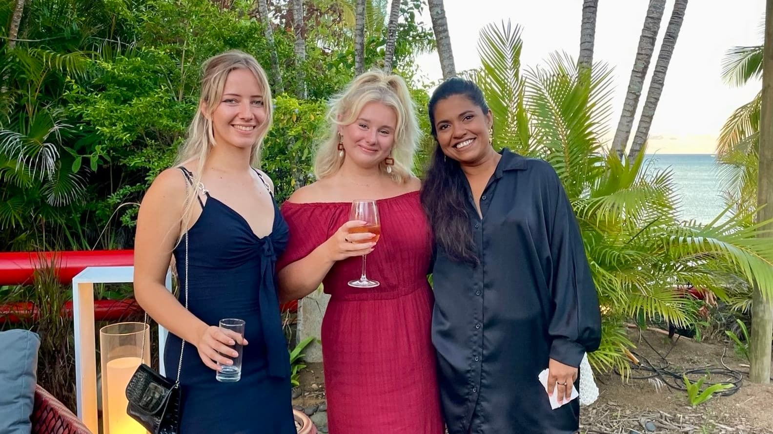 Three women smiling, standing outdoors near tropical plants, one holding a drink. They're dressed in elegant evening wear.
