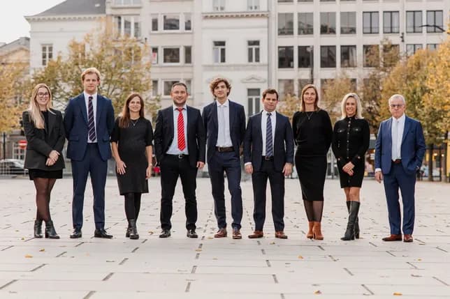 A group of nine people in business attire standing outdoors on a plaza, with buildings and trees in the background.