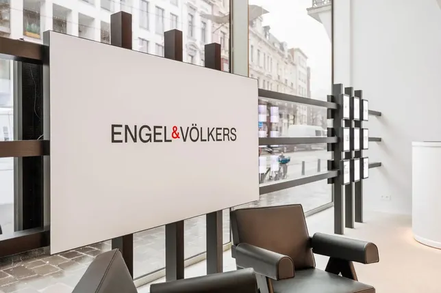 Modern real estate office interior with "ENGEL & VÖLKERS" logo on a wall, chairs, large window, and street view outside.
