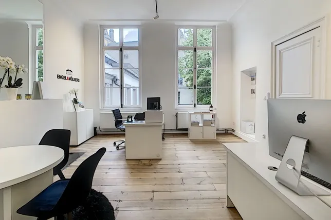 Modern office space with wooden floors, large windows, and minimalist furniture, including desks, chairs, and computers, under bright natural light.