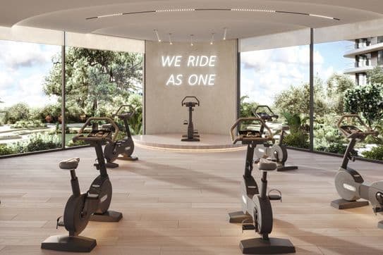 A modern indoor cycling studio with several stationary bikes, large windows, and a sign reading "WE RIDE AS ONE" on the wall.