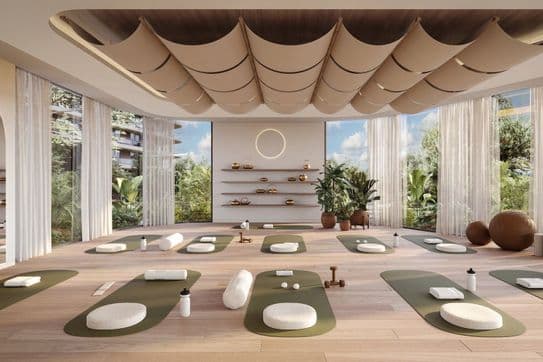 A serene yoga studio with green mats, cushions, and bolsters on a wooden floor. Large windows reveal lush greenery outside.