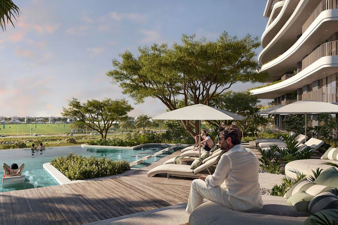 A man in white sits by a modern pool with sun loungers and umbrellas, surrounded by lush greenery and a view of open fields.