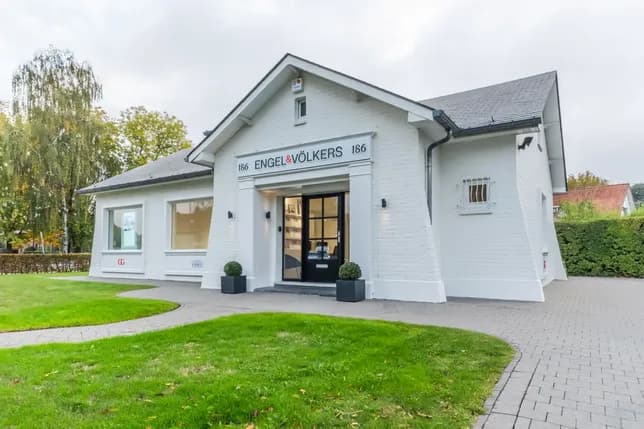 A white brick building with "Engel & Völkers" signage, black door, two potted plants, and a paved walkway, surrounded by greenery.