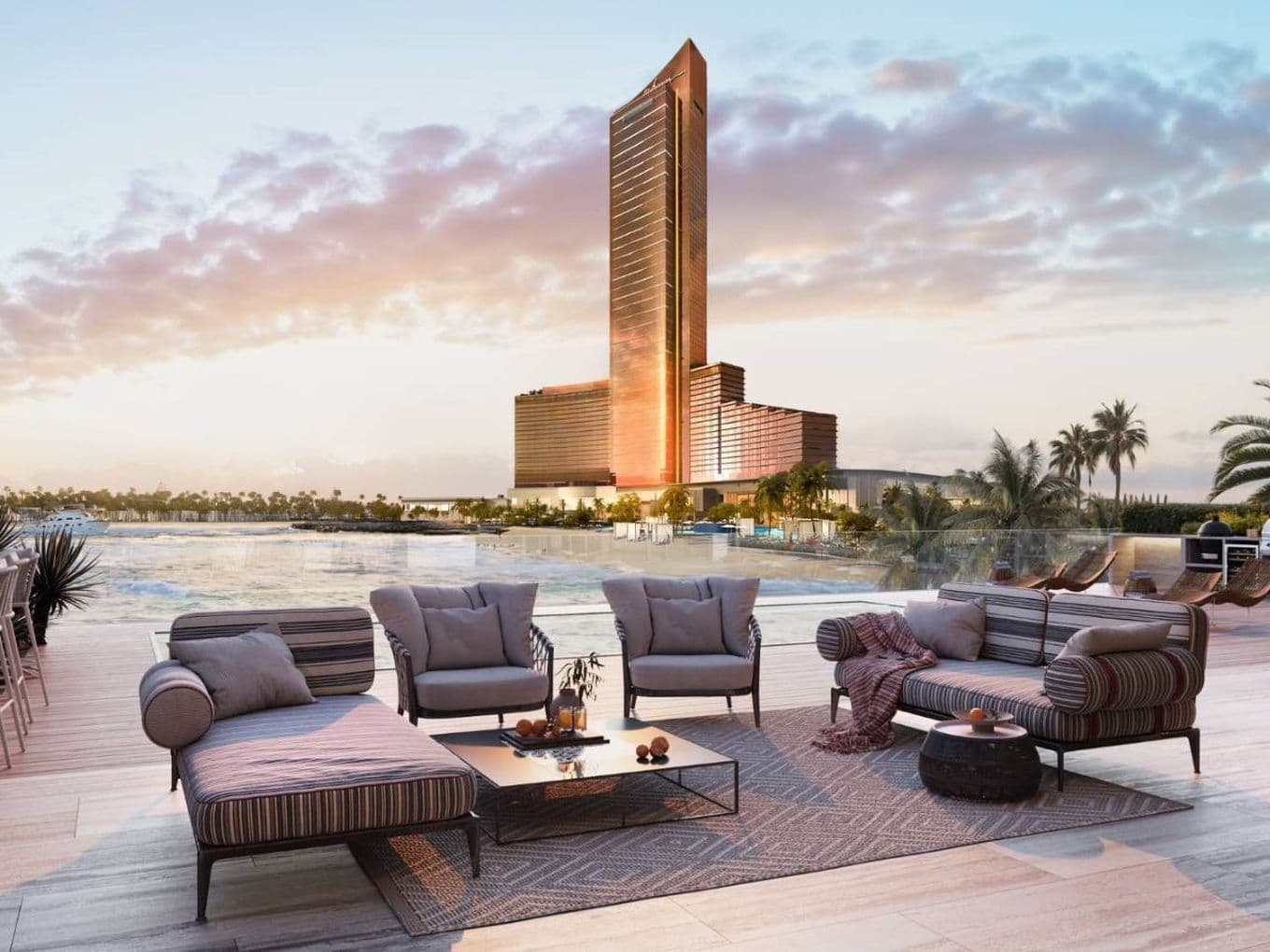 Luxurious outdoor seating area with modern furniture overlooking a large pool and a tall, illuminated building against a colorful sunset sky.