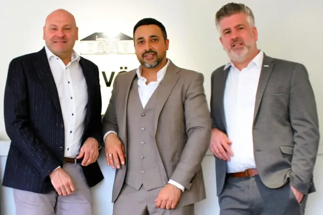 Three men in suits pose confidently against a white wall with a logo. They display a professional demeanor and business attire.