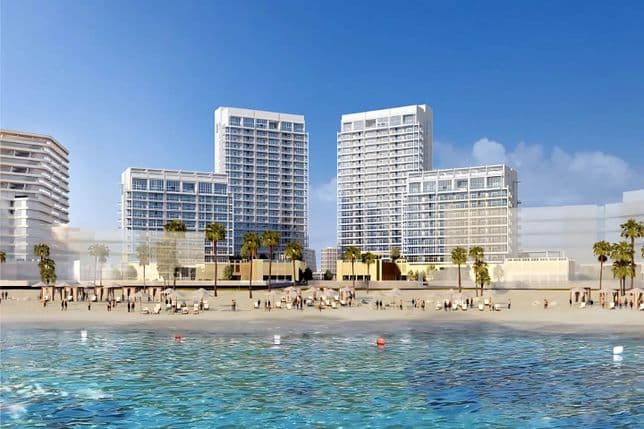 Beachfront view of modern high-rise buildings with palm trees, a sandy beach, and people enjoying the sunny day by the clear blue water.