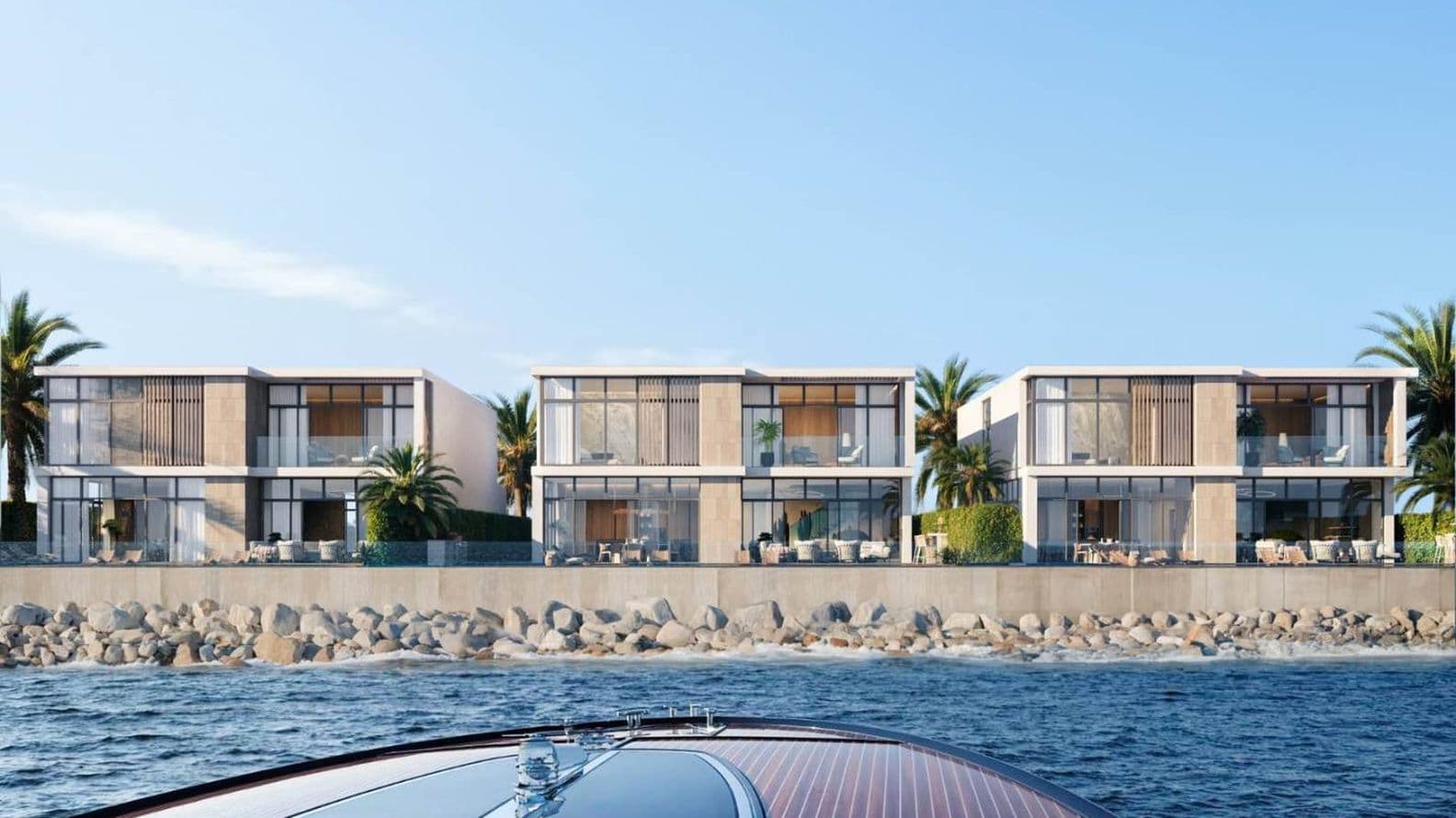 A row of modern beachfront villas with large windows, palm trees, and a stone seawall, viewed from a boat on the water.