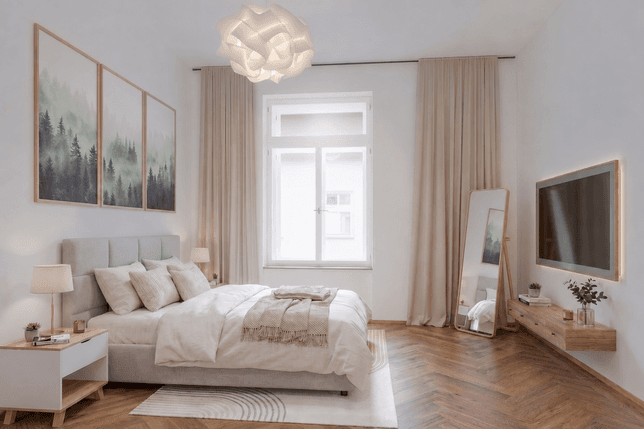 Spacious bedroom with herringbone wooden flooring. A double bed with an upholstered headboard, light bedding, and two bedside tables with lamps is positioned along the left wall. Above the bed hangs a three-panel artwork depicting a misty forest. Opposite the bed, a wall-mounted television is installed above a floating wooden cabinet. A full-length mirror in a wooden frame stands beside the window dressed with beige curtains.