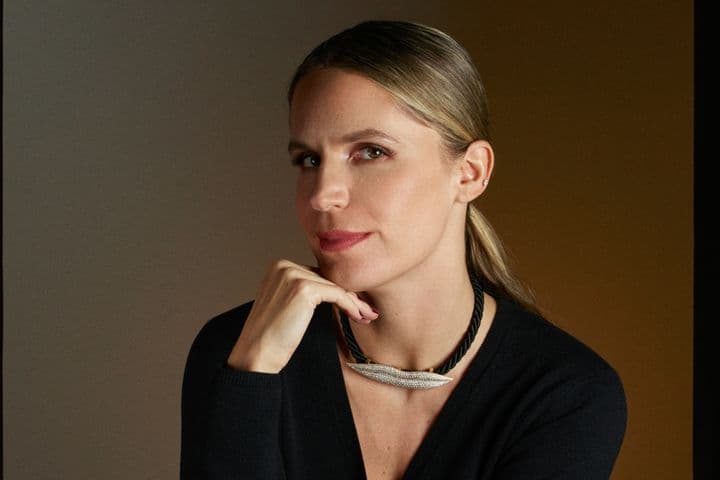 Μια ξανθιά γυναίκα με μαύρο πουλόβερ ποζάρει με το χέρι στο πηγούνι της. Φοράει ένα μαύρο σχοινένιο κολιέ με ένα ασημένιο μενταγιόν.