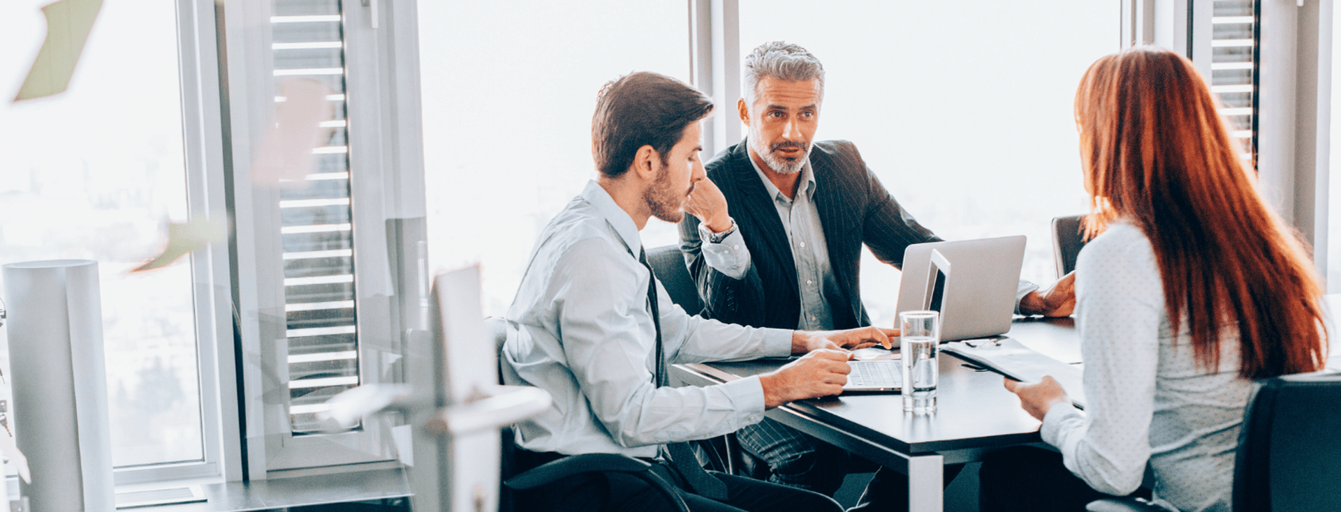 Three business people meet at a table with laptops and papers in a bright office.
