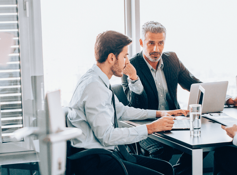Three business people meet at a table with laptops and papers in a bright office.