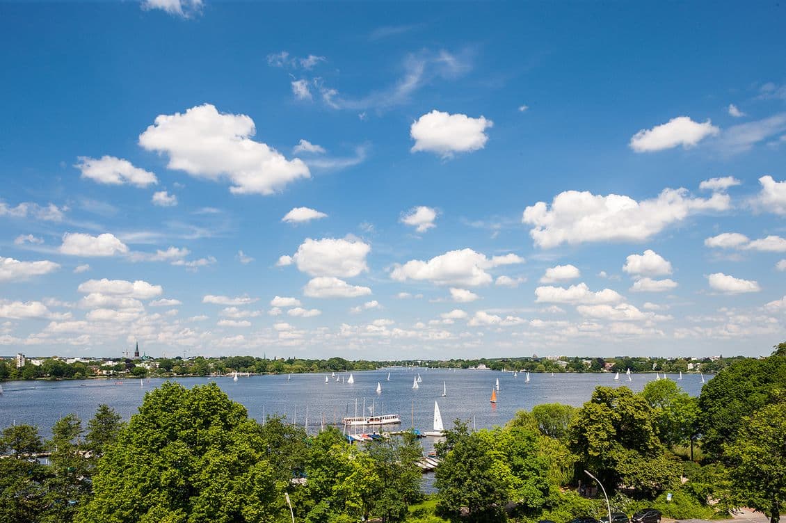 A scenic view of a lake with sailboats, surrounded by lush green trees under a blue sky with scattered clouds.