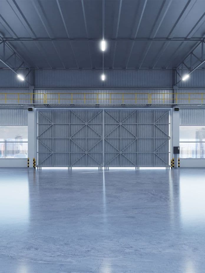 Spacious, empty industrial warehouse with large metal doors, high ceiling, and bright overhead lights illuminating the polished concrete floor.