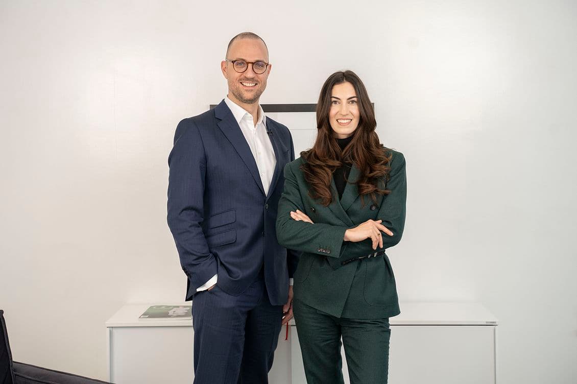 Two professionals in suits smiling, standing in an office with a white background.