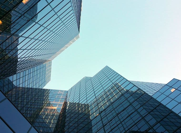 Upward view of modern glass skyscrapers reflecting the sky, creating geometric patterns against a clear blue background.