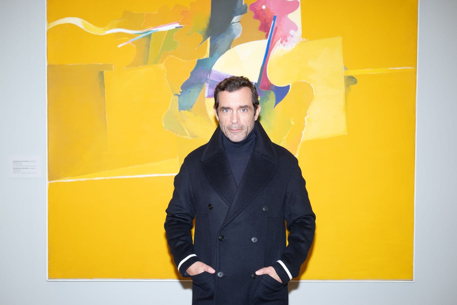 Ένας άνδρας με ένα μπλε ναυτικό παλτό στέκεται μπροστά σε έναν μεγάλο, αφηρημένο πίνακα με κίτρινες, γκρι και ροζ αποχρώσεις.