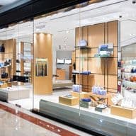 A modern retail store with glass walls displays various handbags and accessories on shelves and stands, with bright lighting highlighting the products.