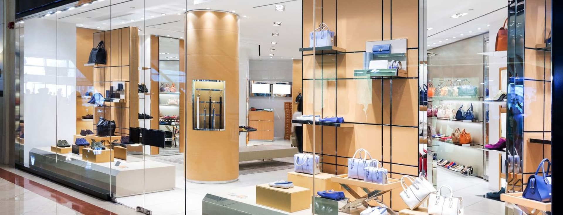 A modern retail store with glass walls displays various handbags and accessories on shelves and stands, with bright lighting highlighting the products.