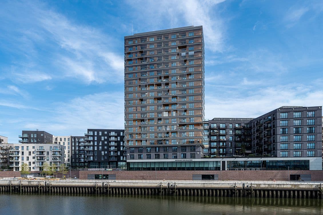 Tall modern apartment building by a waterfront, surrounded by smaller buildings, under a blue sky with scattered clouds.