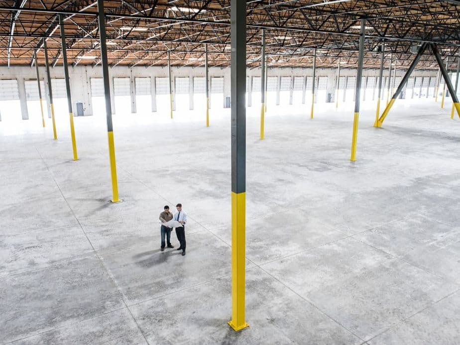 Zwei Personen stehen in einer großen, leeren Lagerhalle mit hohen Decken, umgeben von gelben Säulen und mehreren Garagentoren.