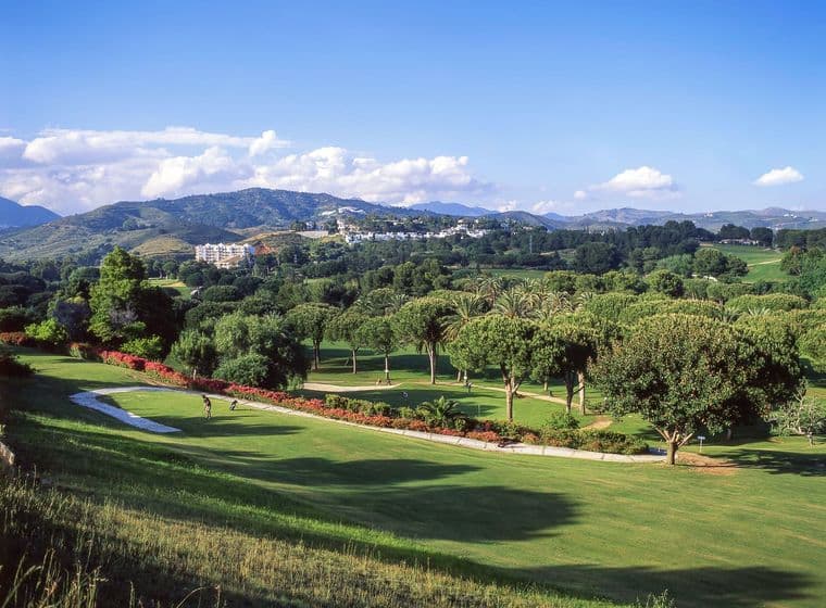Ein malerischer Golfplatz mit sanft geschwungenen grünen Hügeln, Bäumen und fernen Bergen unter einem klaren blauen Himmel.