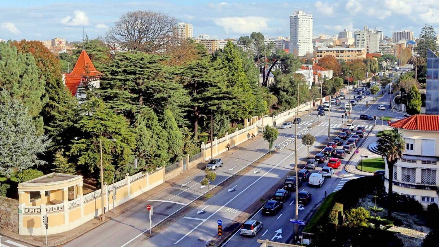 Aerial view of a busy city street lined with trees and buildings, with cars in traffic and a clear sky above.
