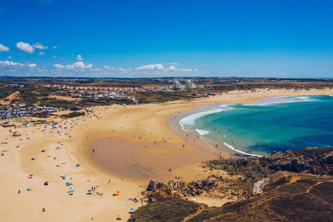 Aerial view of a sandy beach with turquoise waves, scattered beachgoers, colorful umbrellas, and a distant town under a clear blue sky.