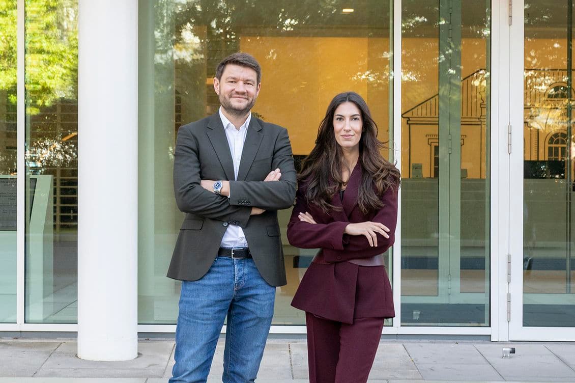 Two people stand confidently with arms crossed in front of a modern glass building. The man wears a blazer and jeans; the woman wears a maroon suit.