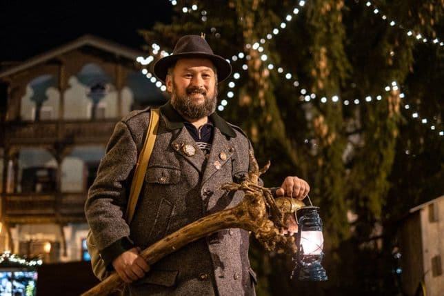 Ein Mann in traditioneller Tracht hält eine Laterne und einen geschnitzten Wanderstock, im Hintergrund weihnachtliche Beleuchtung.
