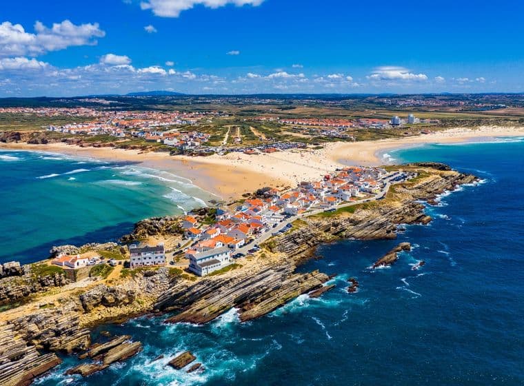 Luftaufnahme einer Küstenstadt mit rotbedachten Gebäuden, Sandstränden, felsigen Klippen und türkisfarbenem Ozean unter einem klaren blauen Himmel.