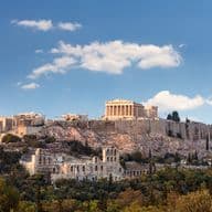 The Acropolis in Athens, Greece, with the Parthenon atop a rocky hill, under a blue sky with scattered clouds. Surrounding greenery visible.