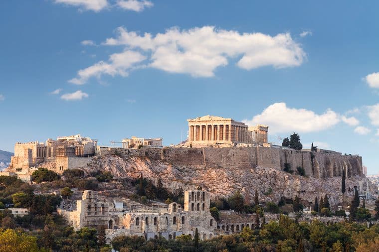 The Acropolis in Athens, Greece, with the Parthenon atop a rocky hill, under a blue sky with scattered clouds. Surrounding greenery visible.