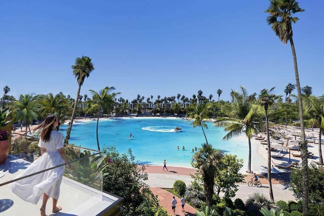Woman in a white dress overlooks a tropical lagoon with palm trees, lounge chairs, and a person jet-skiing under a clear blue sky.