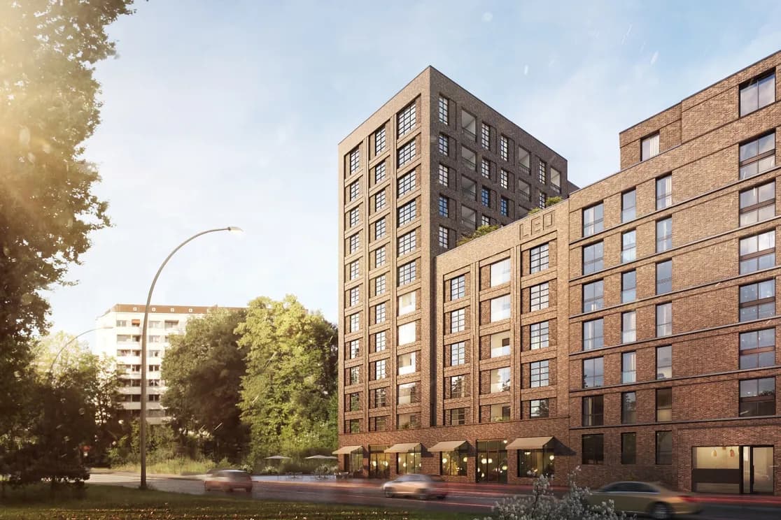 Wohnkomplex aus Backstein mit einer hohen Fassade im Rastermuster neben einem niedrigeren Backsteinblock; geschwungene Straßenlaterne, vorbeifahrende Autos, Bäume und ein strahlend blauer Himmel.